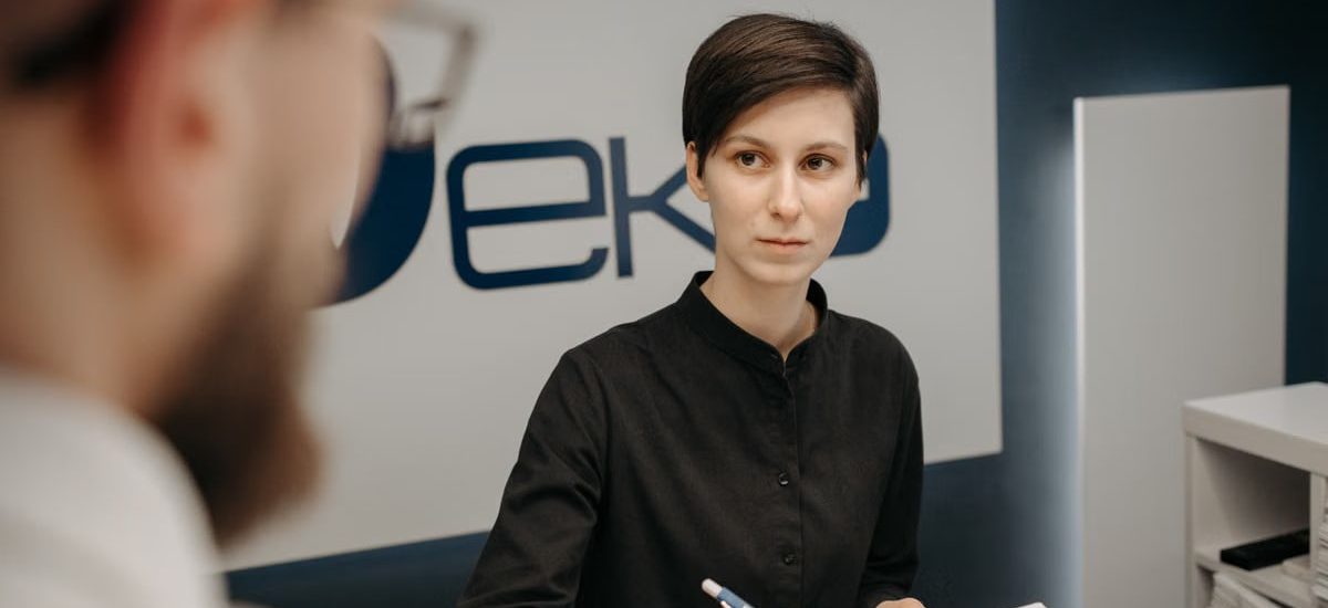 A receptionist at a clinic desk writing notes during a consultation.
